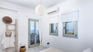 A minimalist bedroom with a large bed, a hanging light fixture, a view of the sea through a window, and a balcony door. - Villa Vinea (Santorini)
