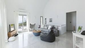 A spacious living area with a sofa, coffee table, and bean bag, a balcony with a chair, and a white wall with a framed picture. - Villa Vinea (Santorini)