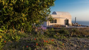 A small, white building with a dome roof, surrounded by greenery and flowers, overlooking a body of water. - Villa Vinea (Santorini)