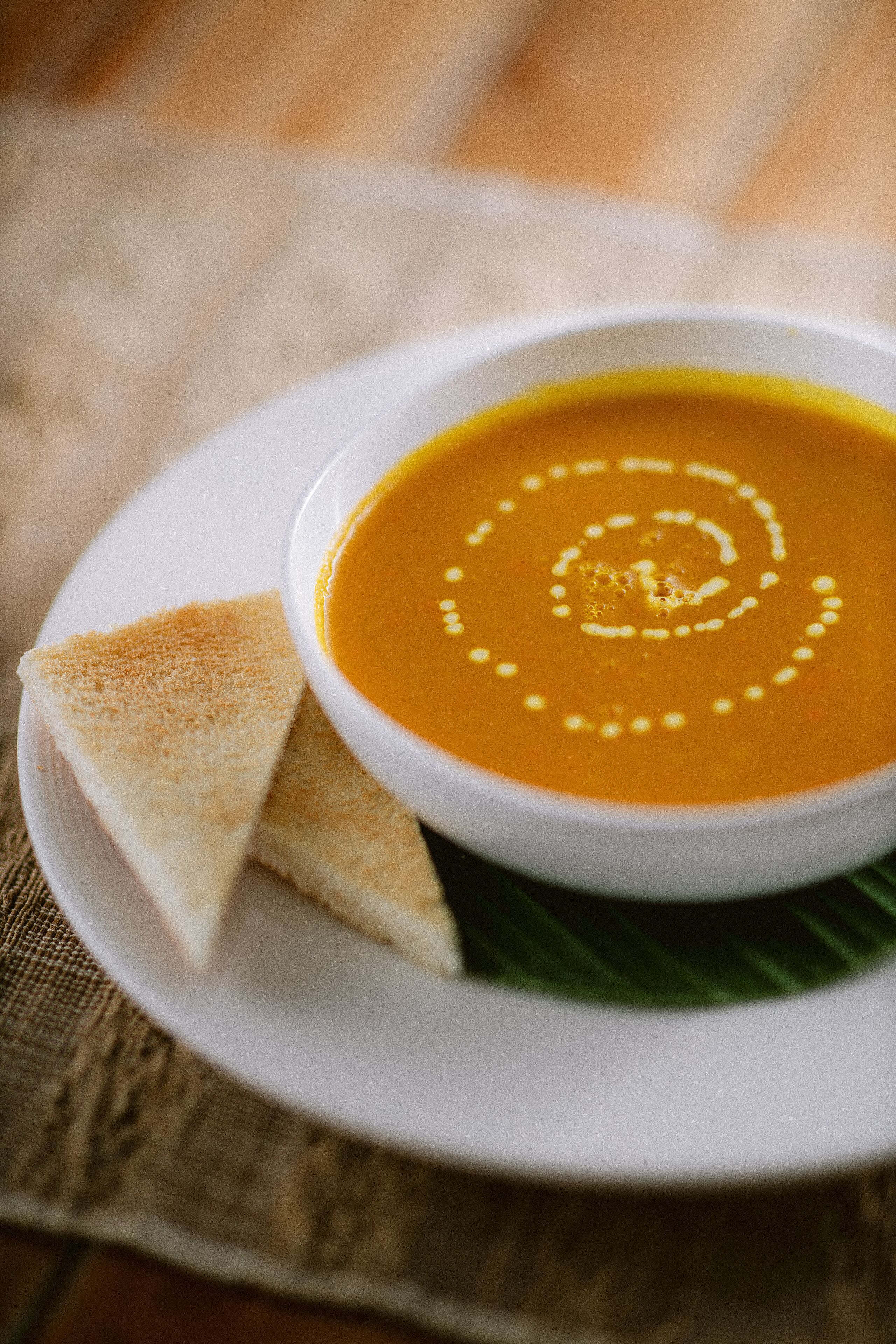 A bowl of soup on a white plate with two slices of bread beside it, placed on a table with a woven placemat.