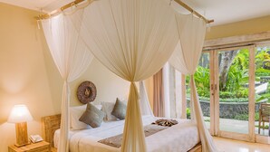 Een slaapkamer met hemelbed, nachtkastje met lamp en uitzicht op een tropische tuin door de glazen deur.