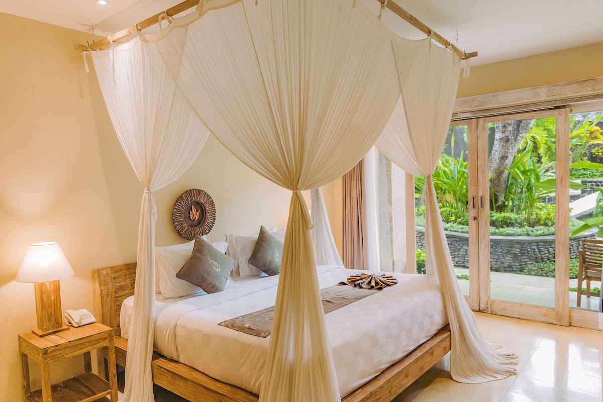 Een slaapkamer met hemelbed, nachtkastje met lamp en uitzicht op een tropische tuin door de glazen deur.