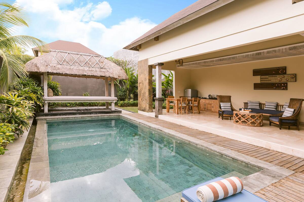 Zwembad met een rieten parasol, houten terras en een buiten zitgedeelte met stoelen en een tafel.