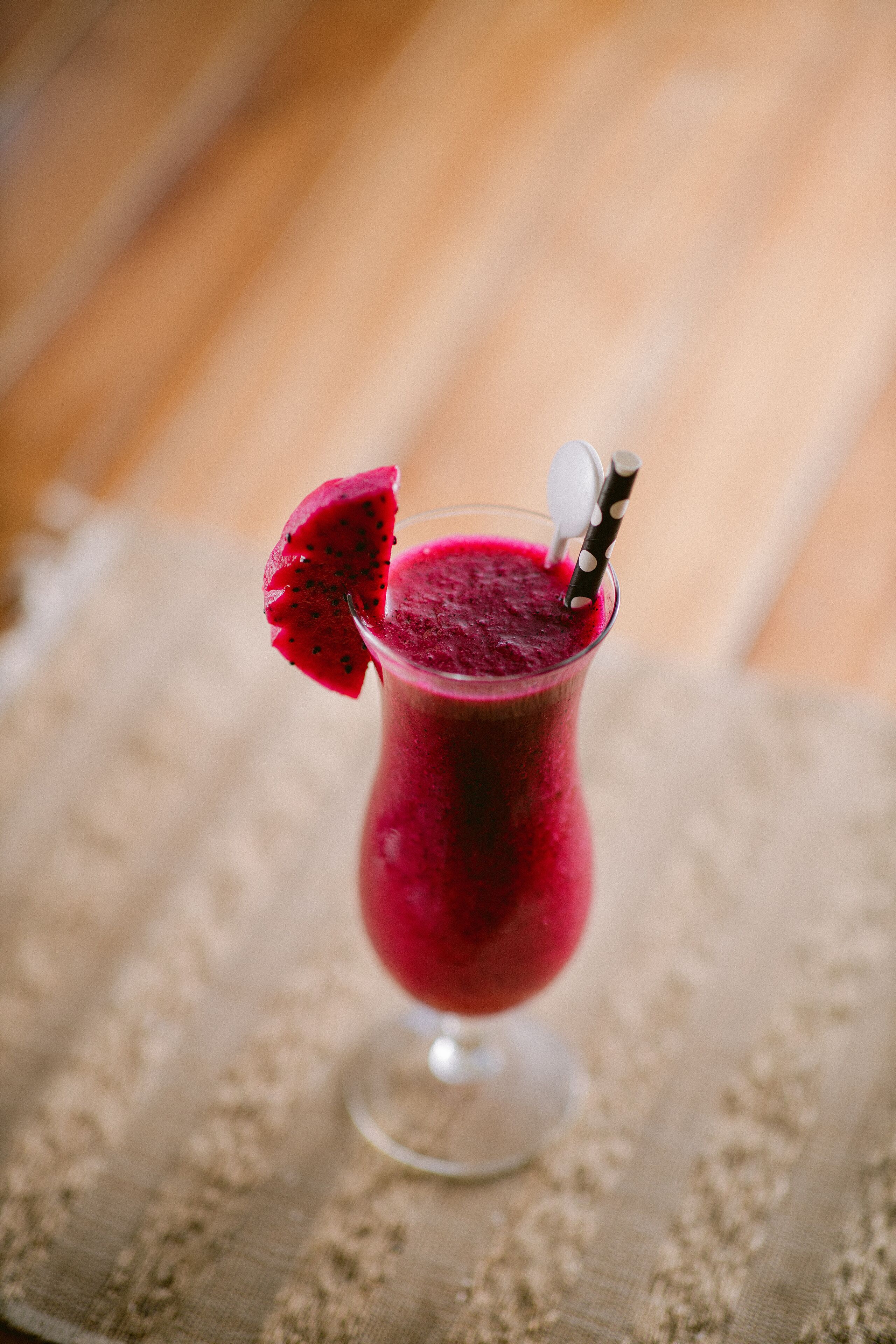 A glass of red smoothie with a dragon fruit slice garnish and a spoon, placed on a textured cloth.