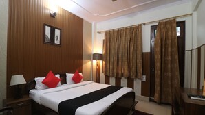 A hotel room with a bed, two bedside tables, a lamp, a wall-mounted light, a framed picture, and a window with curtains. - Hotel Mandakini (Jakholi)
