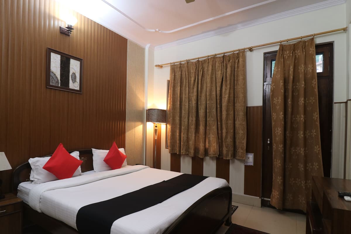 Una habitación de hotel con cama, dos mesitas de noche, una lámpara, luz empotrada en la pared, un cuadro enmarco y una ventana con cortinas.