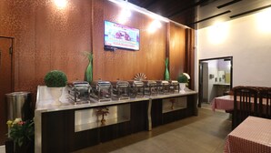 A buffet setup with multiple serving stations, a television mounted on the wall, and a seating area with tables and chairs. - Hotel Mandakini (Jakholi)