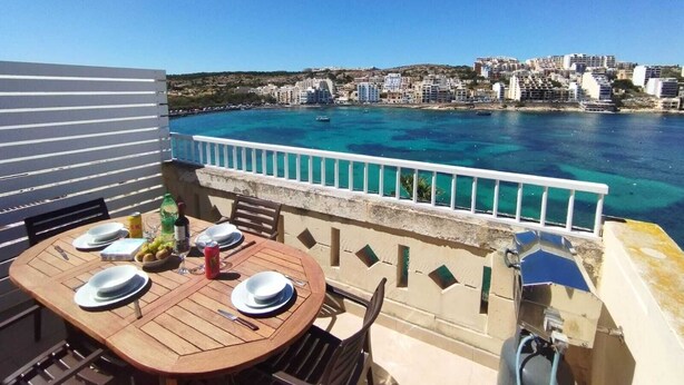 Mesa de madera dispuesta para comer, con platos, cubiertos y una botella, con vistas a un pueblo costero con edificios y un mar azul cristalino.