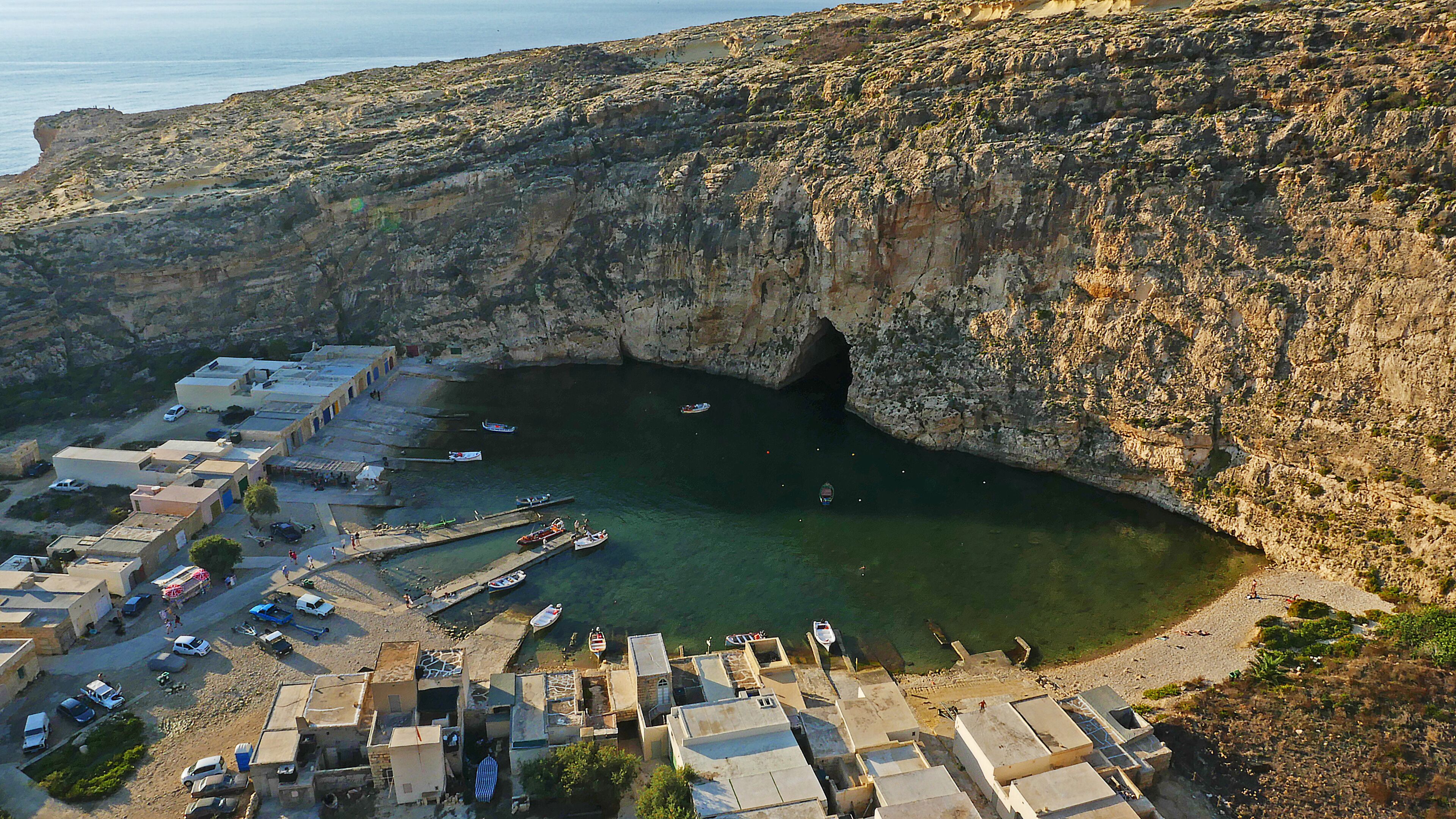 A coastal village with a rocky cliff, a harbor with boats, and buildings with flat roofs.