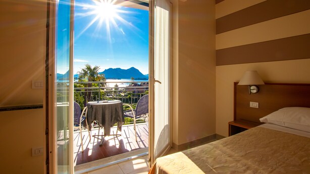 Habitación de hotel con balcón, cama, mesita de noche y vistas a la playa y montañas.