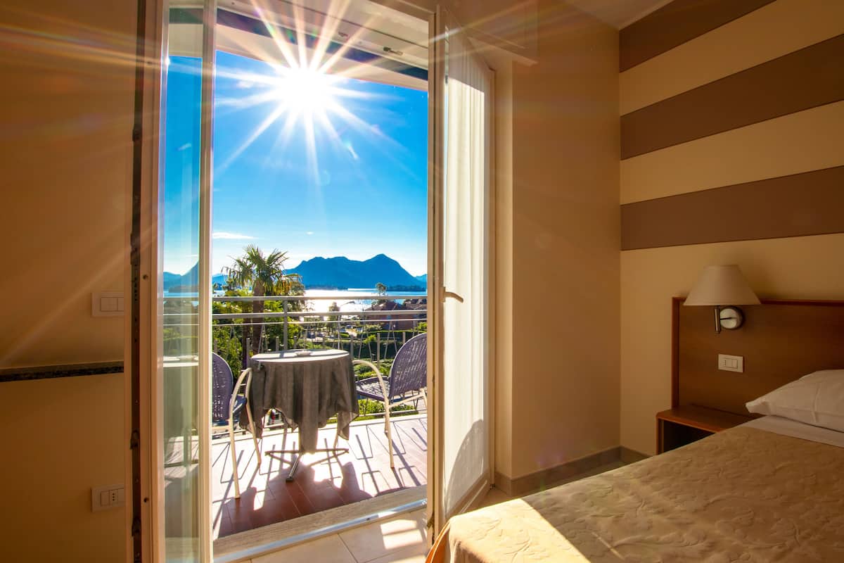 A hotel room with a balcony, a bed, a nightstand, and a view of a beach and mountains.