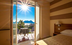 A hotel room with a balcony, a bed, a nightstand, and a view of a beach and mountains. - Hotel Alpi (Baveno)