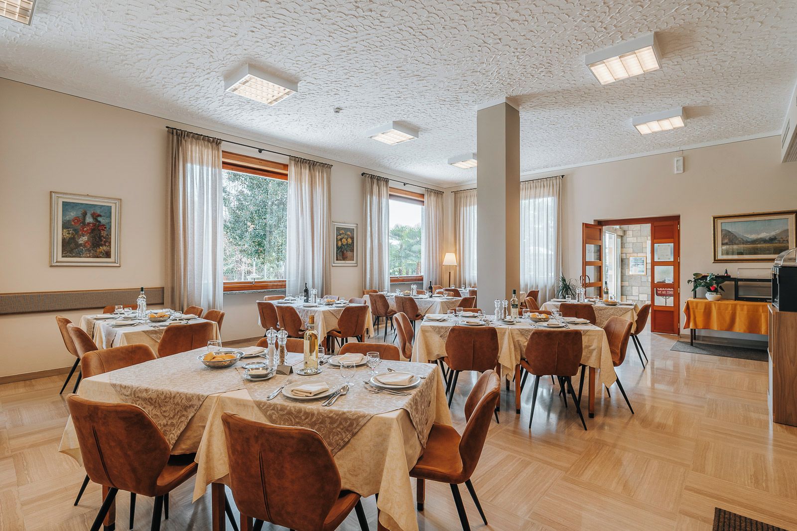 Un comedor con varias mesas y sillas, ventanas amplias con cortinas y vista a zonas verdes.