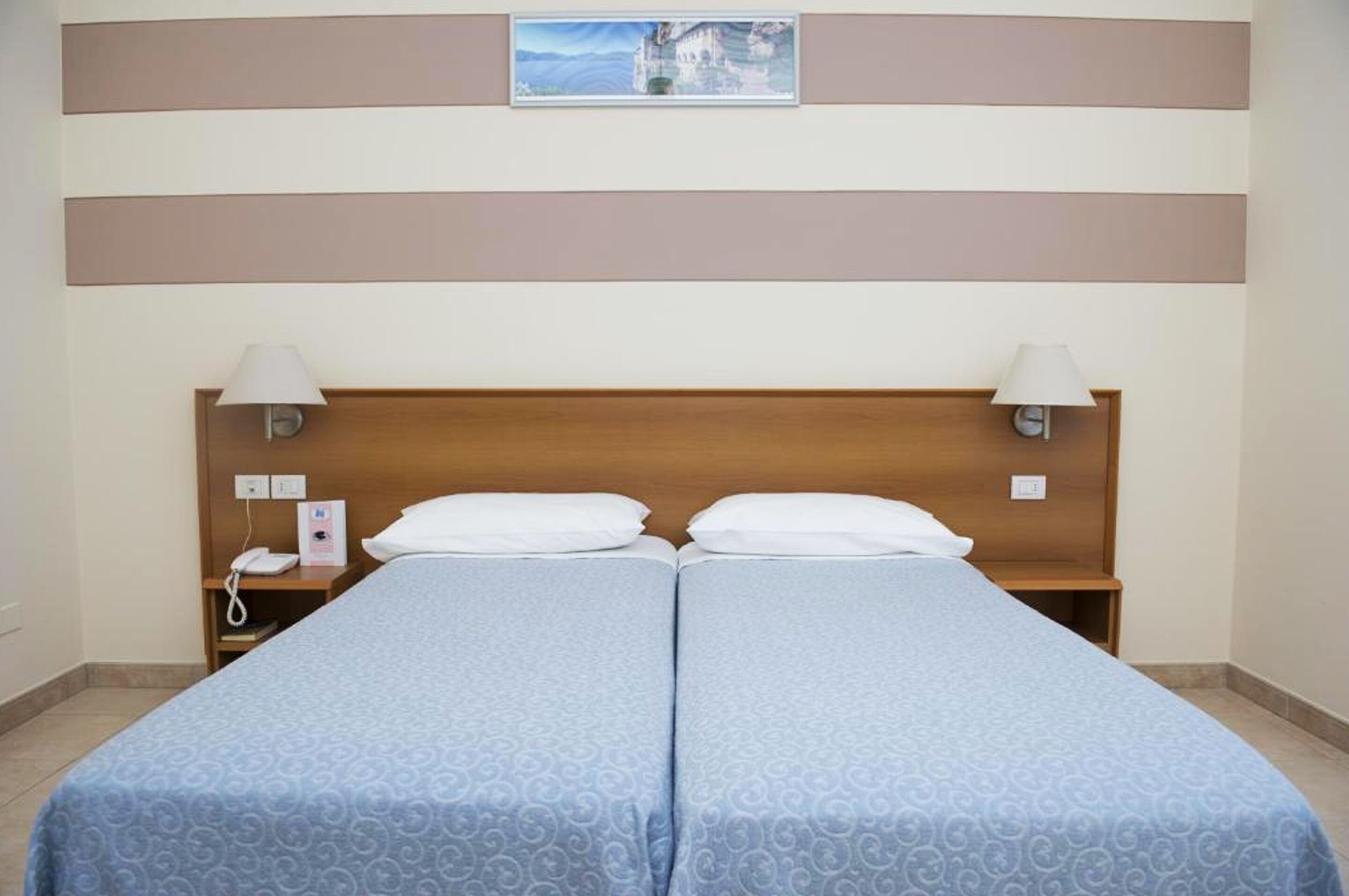 Uma cama de casal com cabeceira de madeira, duas luminárias de cabeceira, uma mesinha de cabeceira com telefone e um quadro emoldurado acima da cama.