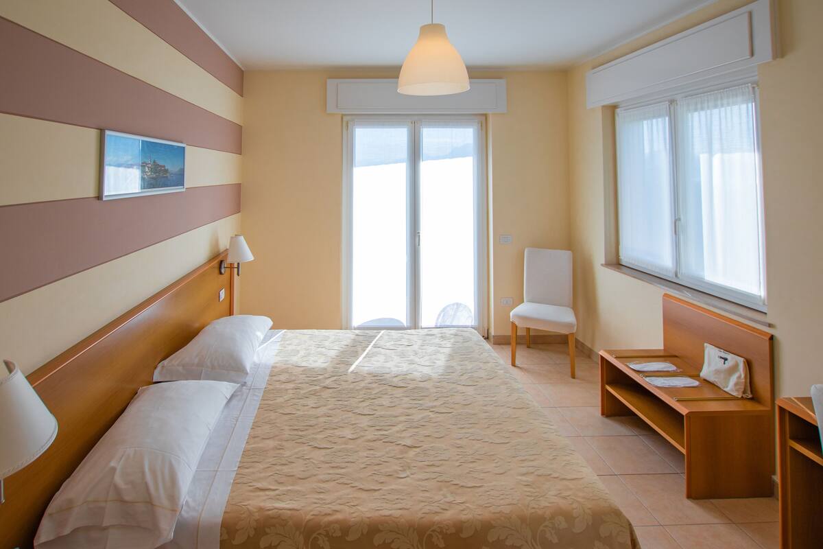 A hotel room with a bed, bedside lamps, a chair, a desk, and a large window with curtains.