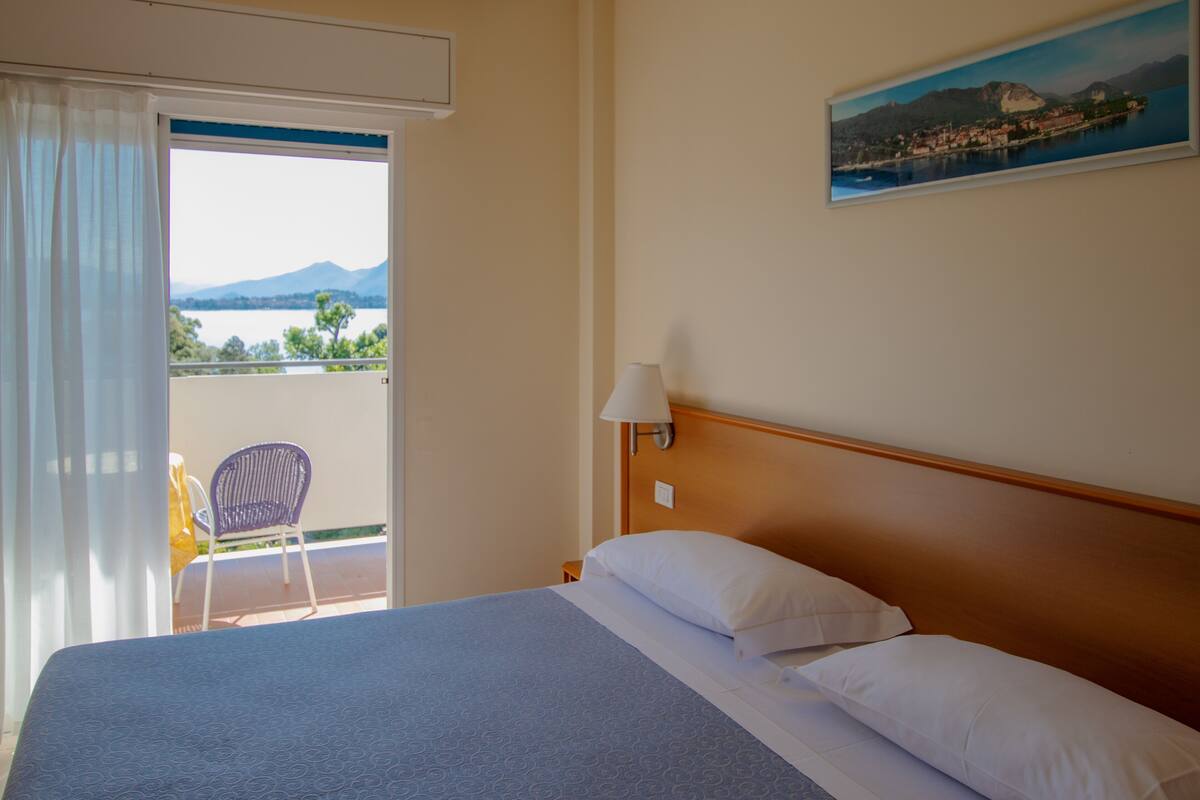 A hotel room with a bed, a chair, a painting, and a view of a lake and mountains.