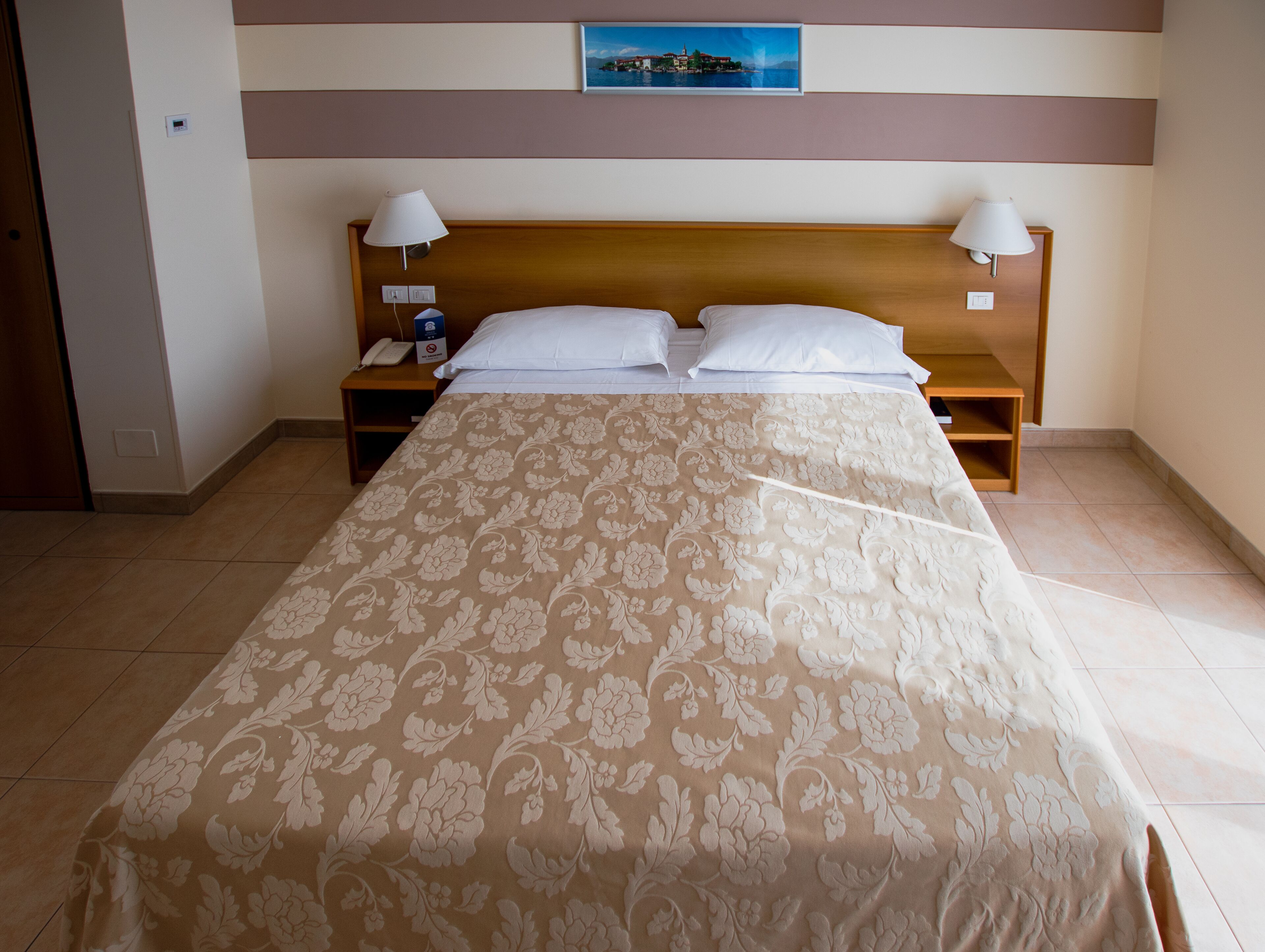 A neatly made bed with a floral bedspread, two bedside lamps, a nightstand with a phone, and a painting above the headboard.