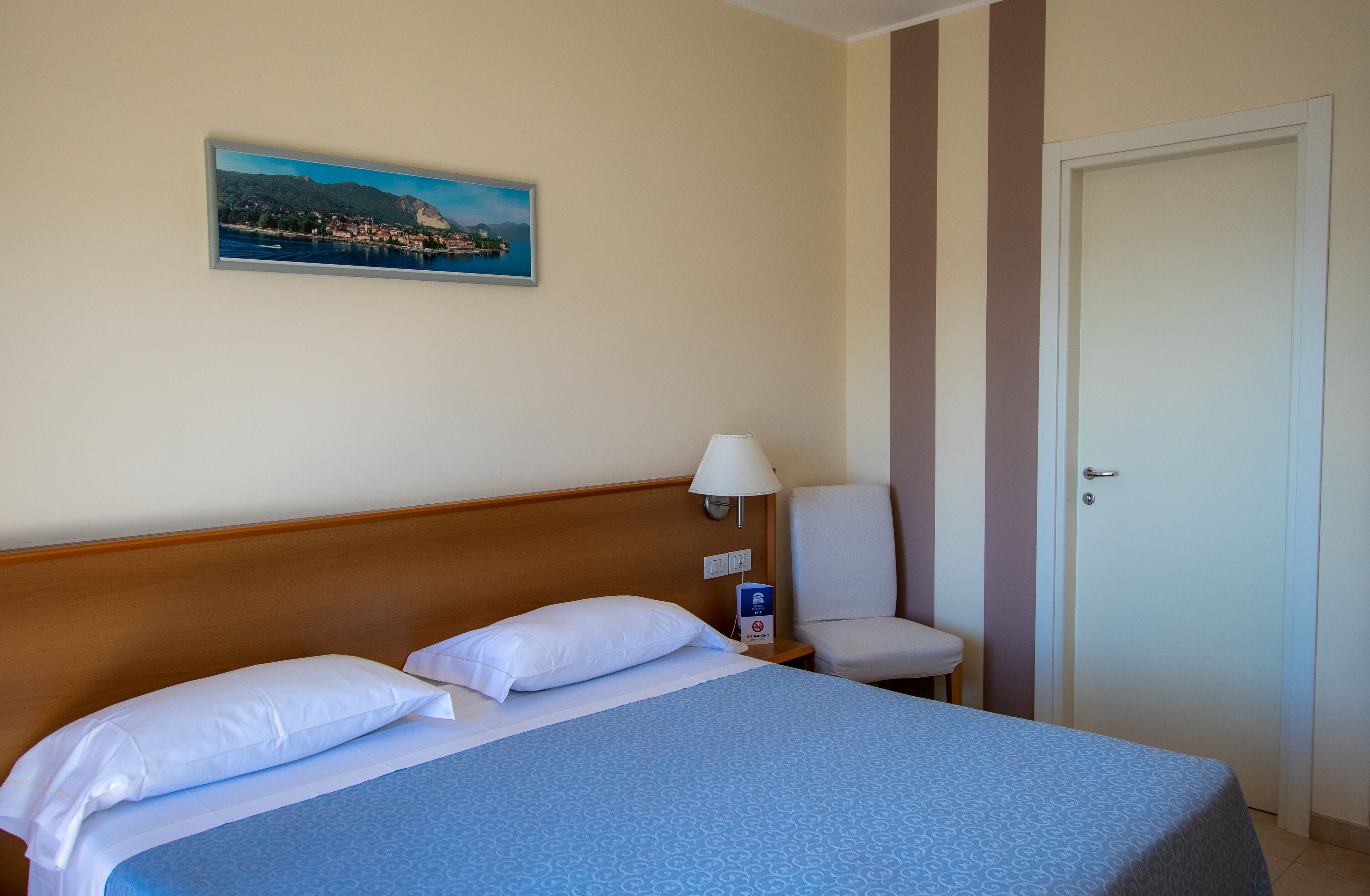 Een hotelkamer met een bed, nachtkastje, lamp, stoel en een deur.