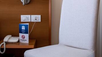 Quarto de hotel com uma poltrona branca, uma mesinha de cabeceira com luminária, um telefone e um aviso de 'Não Fumar'.