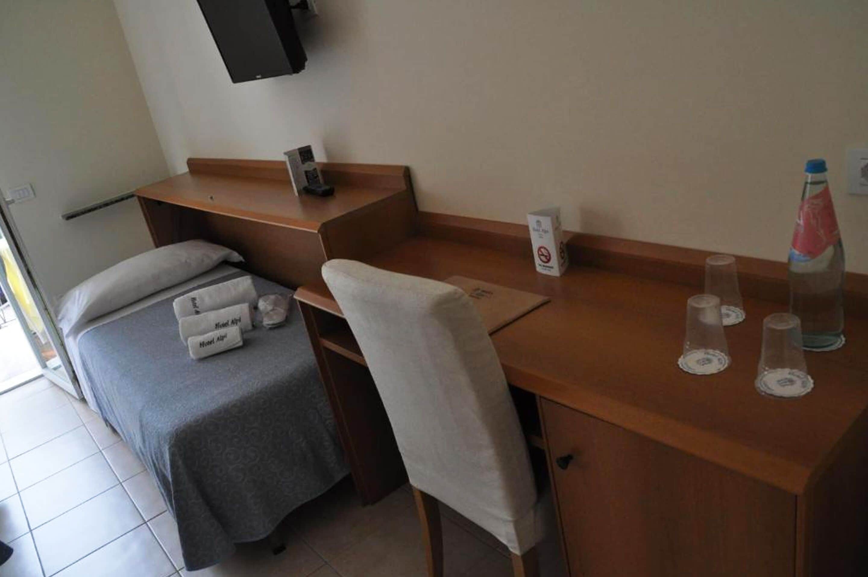 Ein kleiner, ordentlicher Raum mit einem Bett, einem Schreibtisch und einem Stuhl. Auf dem Schreibtisch steht eine Wasserflasche und eine Kiste.
