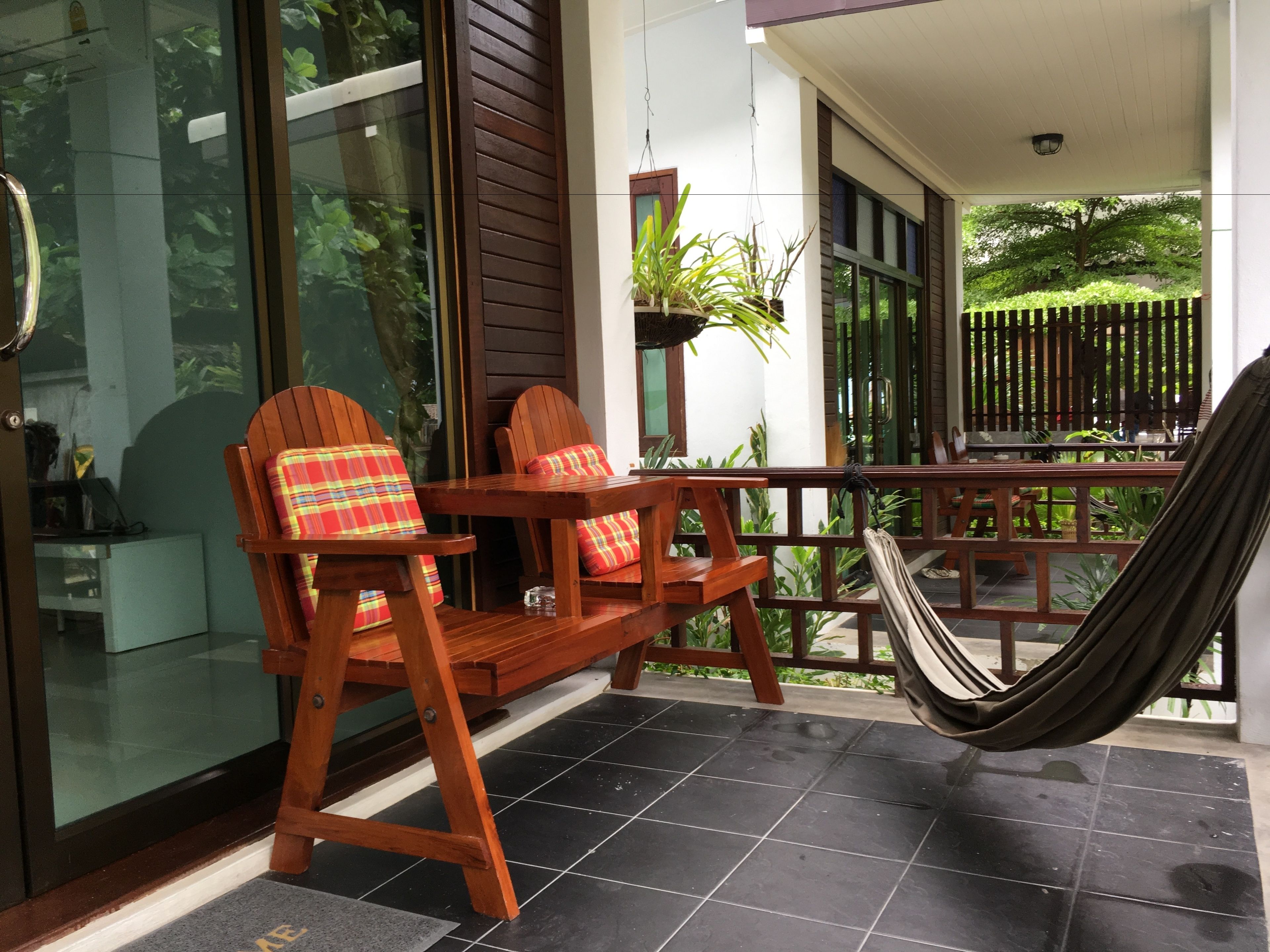 A wooden deck with a hammock, two wooden chairs with plaid cushions, and a glass door leading to a garden.