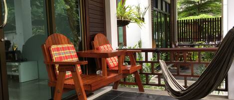 A wooden deck with a hammock, two wooden chairs with plaid cushions, and a glass door leading to a garden.