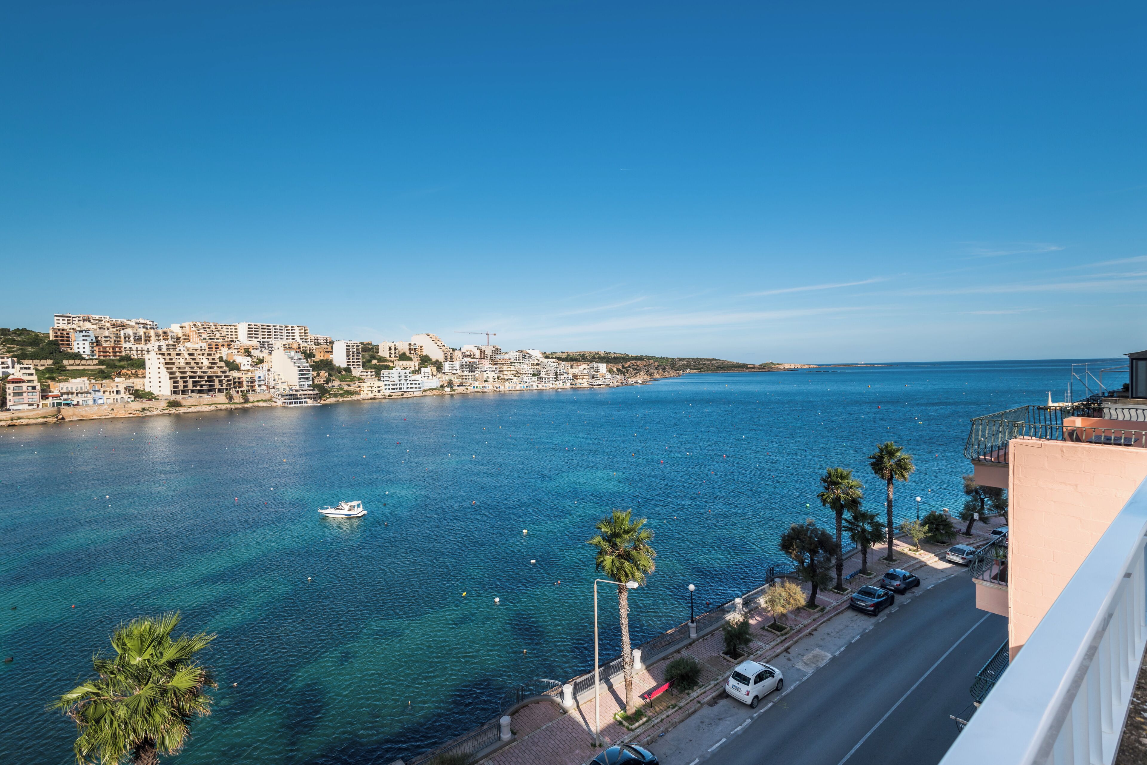 A coastal cityscape with a clear blue sea, a boat on the water, and a road with parked cars.