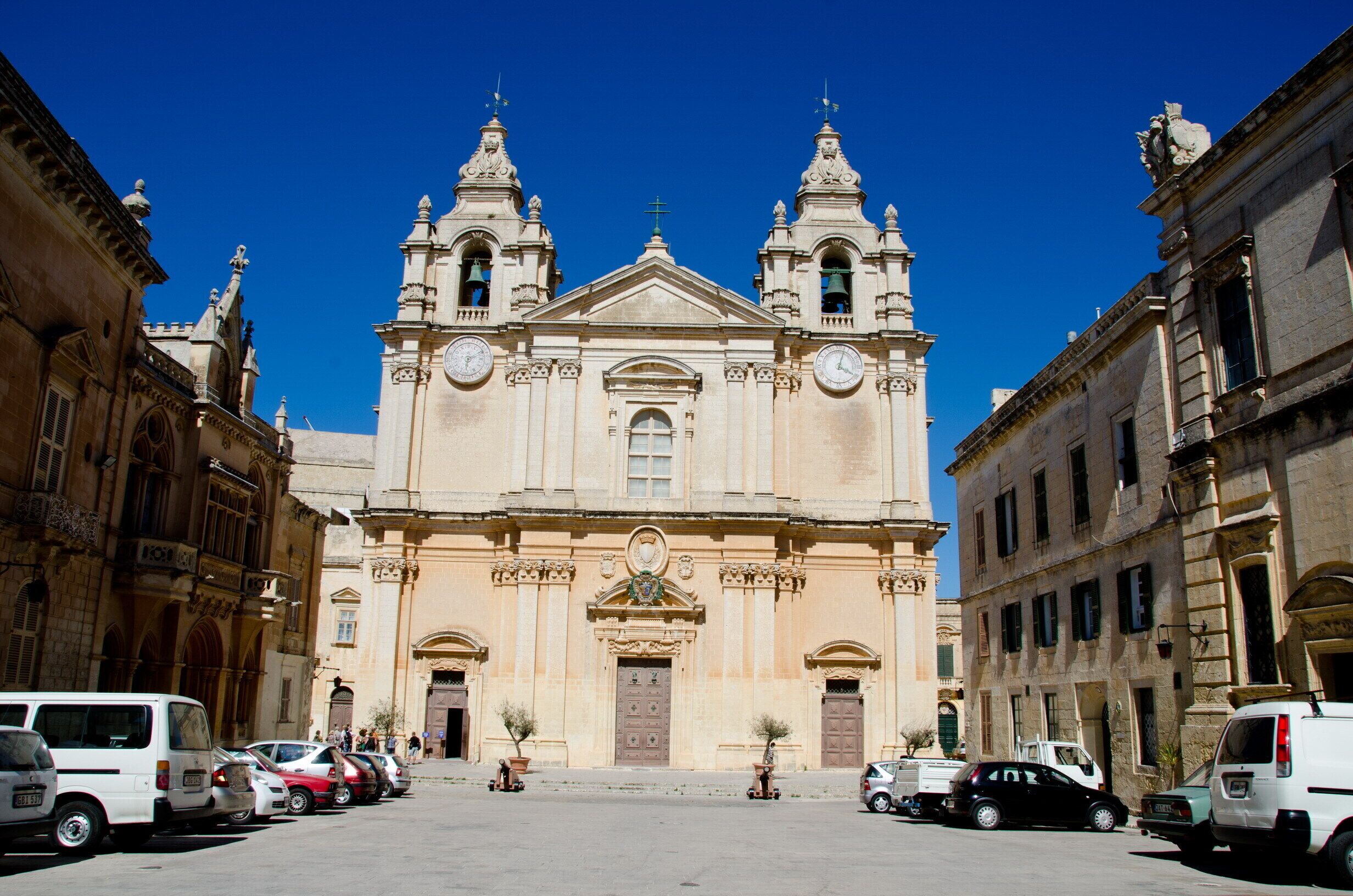 A historic building with a central dome and two bell towers, surrounded by parked cars and other buildings.