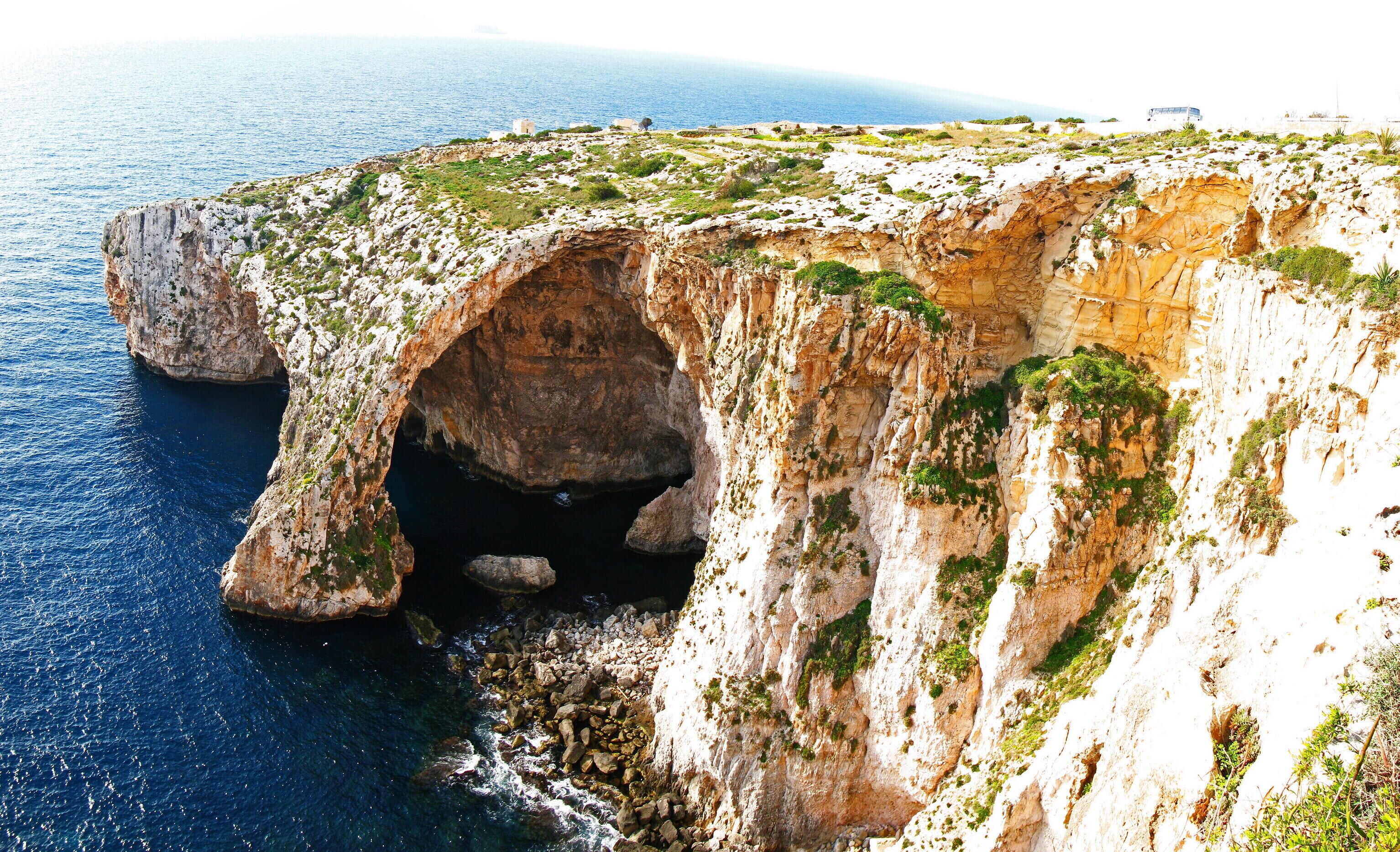 A coastal cliff with a natural cave entrance, rocky terrain, and sparse vegetation.