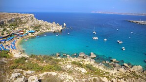 A coastal area with clear turquoise waters, rocky terrain, and a beach with umbrellas. - Blue Harbour 1 by Getaways Malta (St. Paul's Bay)