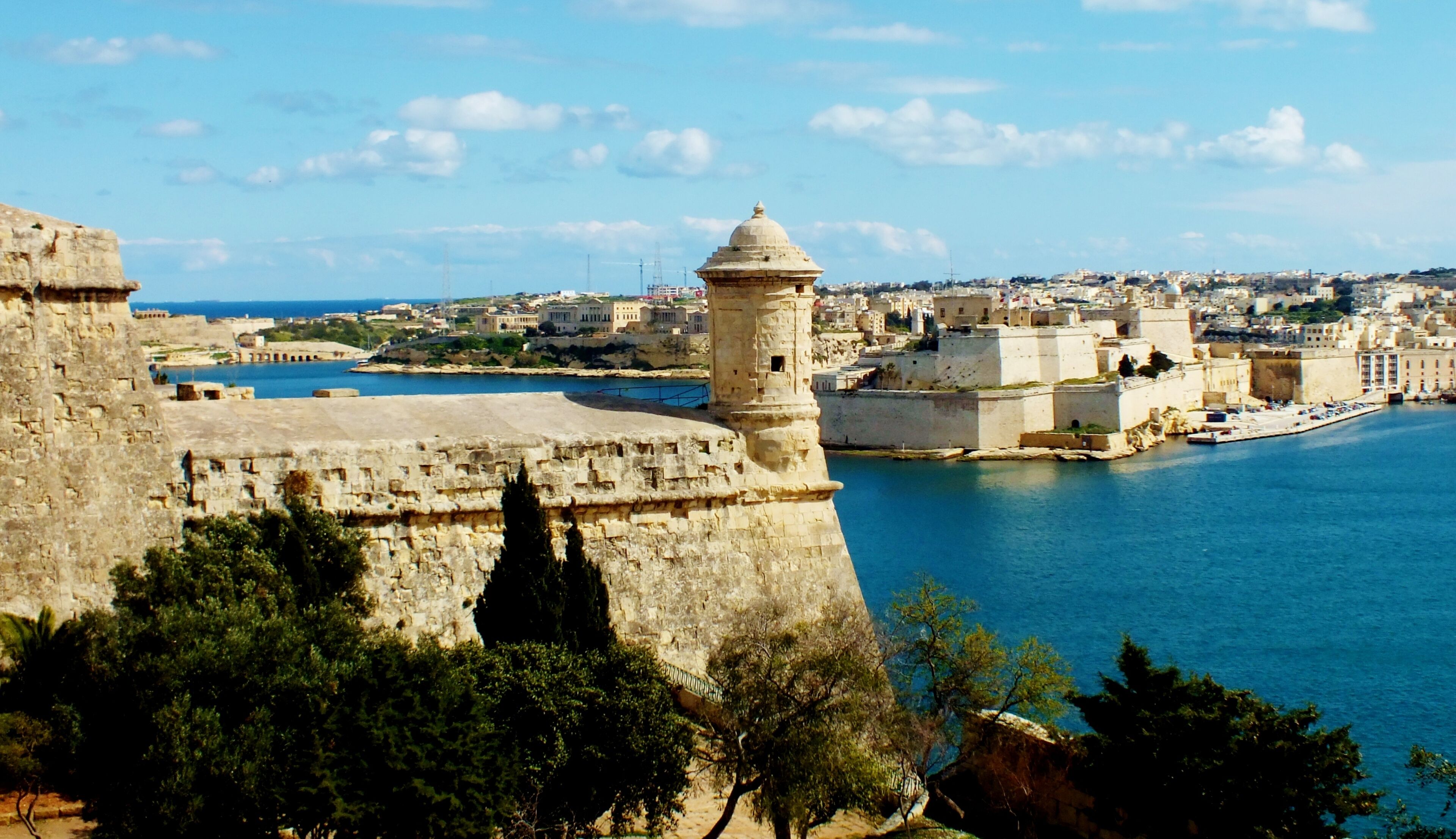 A coastal cityscape with historic stone fortifications, a prominent tower, and a harbor.