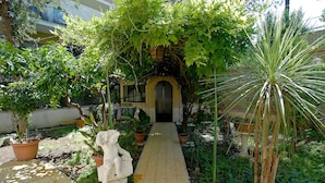 Un chemin de jardin menant à l’entrée d’un bâtiment, bordé de plantes en pot et orné d’une statue.