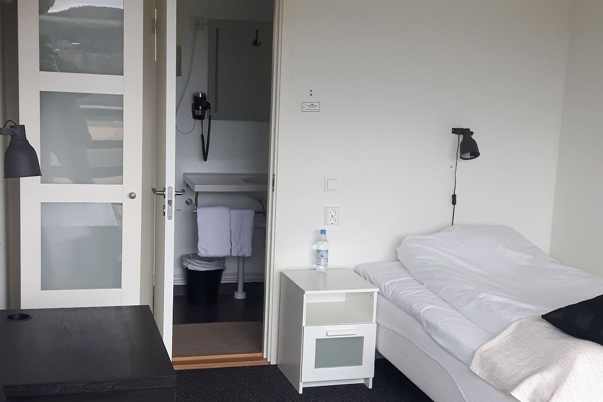 Quarto de hotel com uma cama, escrivaninha, cadeira, mesa de cabeceira, abajur, garrafa de água e banheiro visível através de uma porta aberta.
