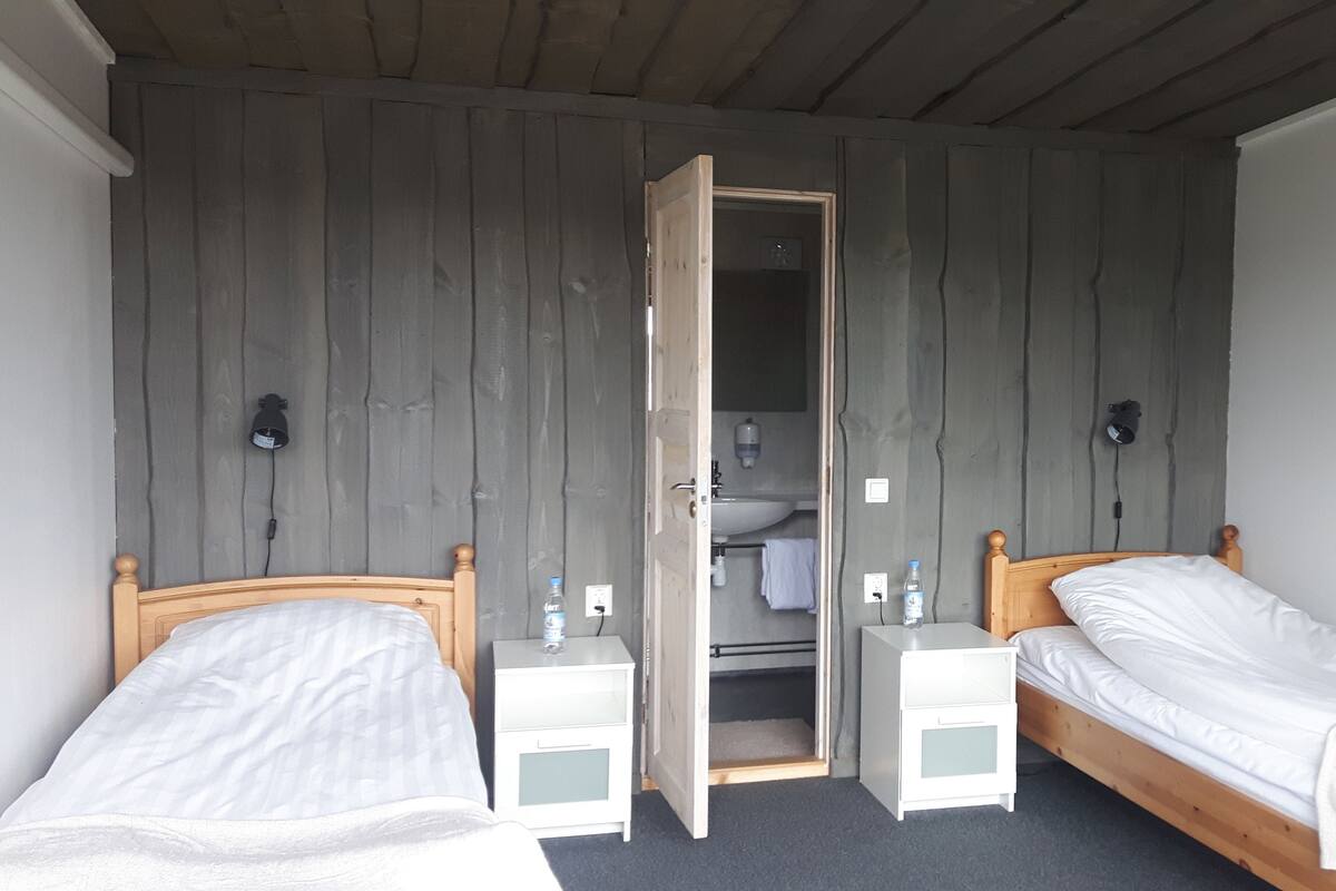 Quarto pequeno e simples com duas camas individuais, um guarda-roupa de madeira, uma mesinha de cabeceira com luminária e um banheiro visível por uma porta aberta.