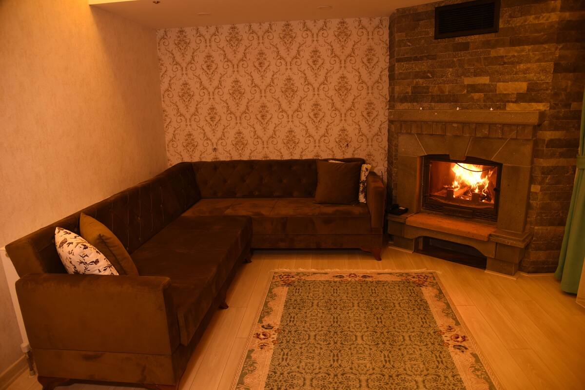 A living room with a brown sectional sofa, a patterned rug, and a stone fireplace with a fire.