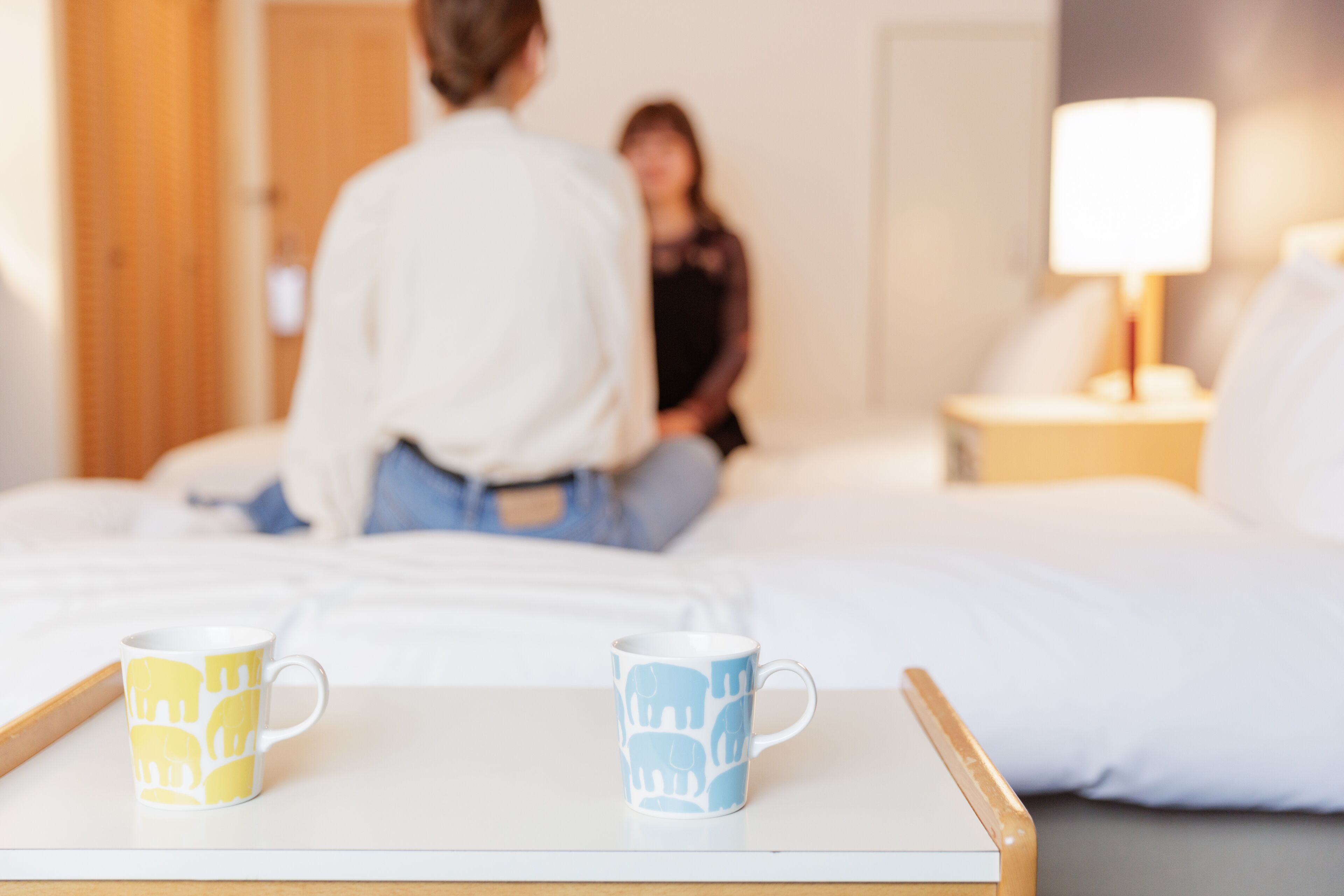 Une chambre d’hôtel avec un lit, une table de chevet sur laquelle sont posées deux tasses et une petite table de nuit avec une lampe.