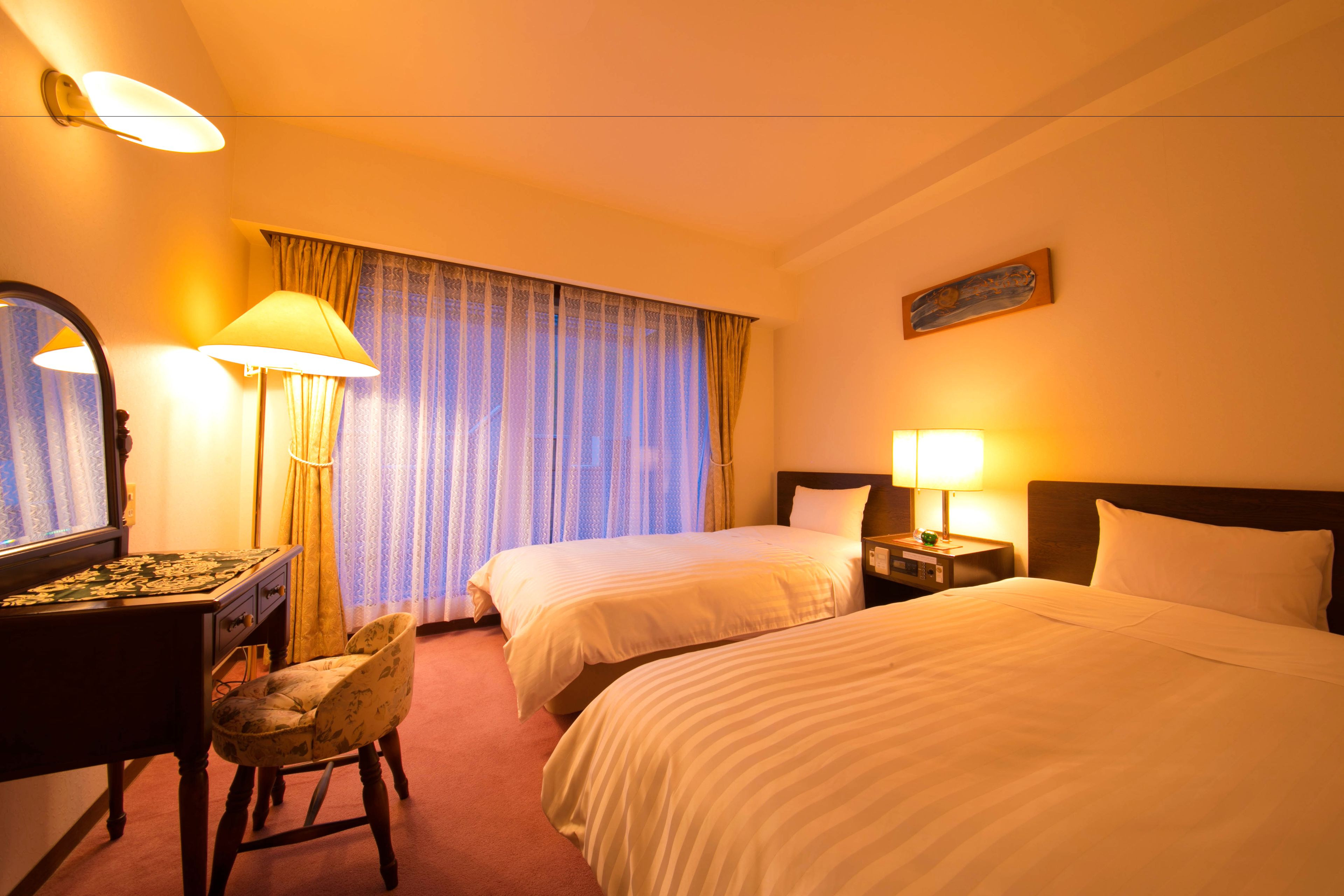 A hotel room with two beds, a desk with a lamp, a chair, a dresser, and a large window with curtains.