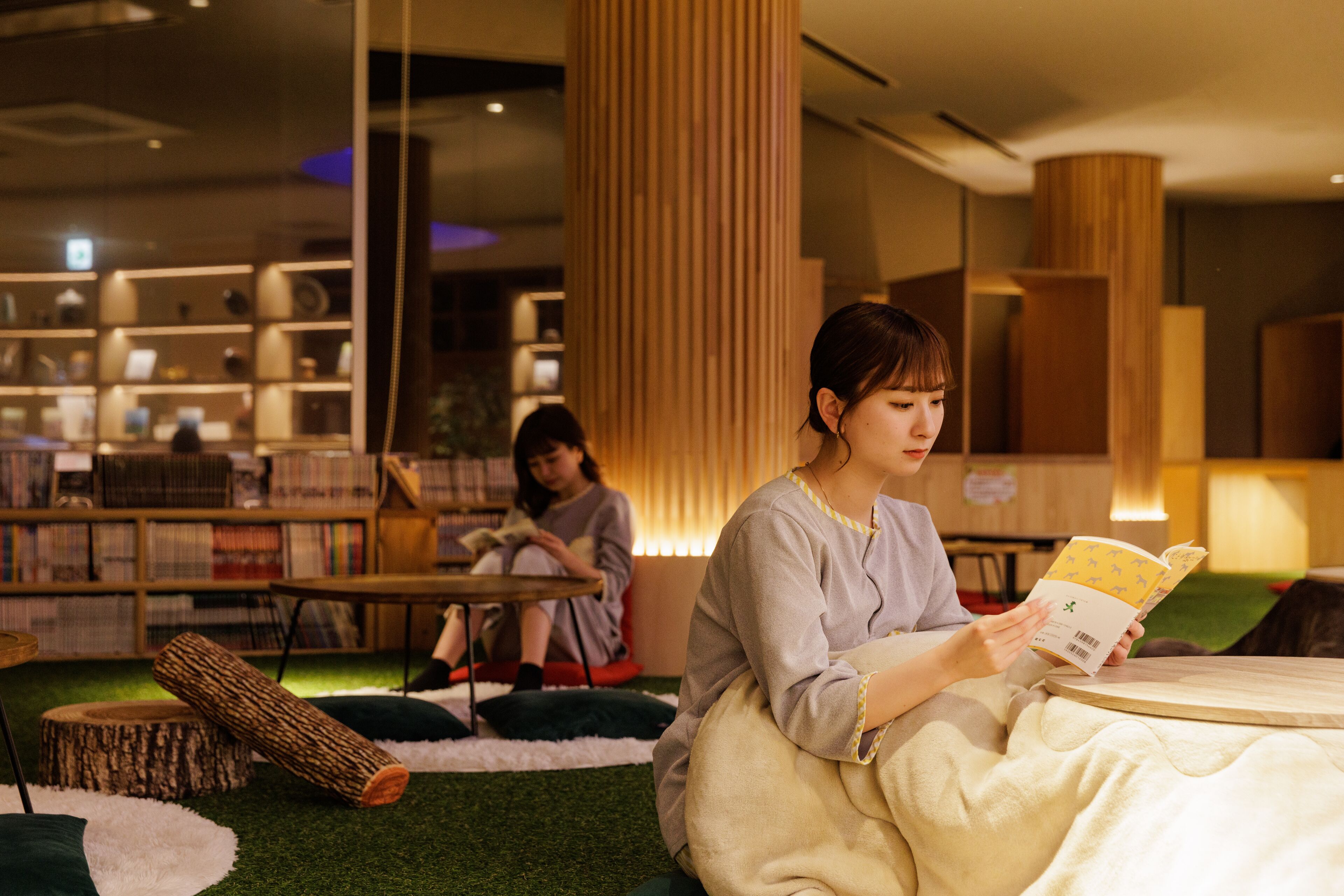 A library setting with bookshelves, a reading area with a person reading a book, and another person sitting in the background.