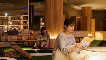 A library setting with bookshelves, a reading area with a person reading a book, and another person sitting in the background.