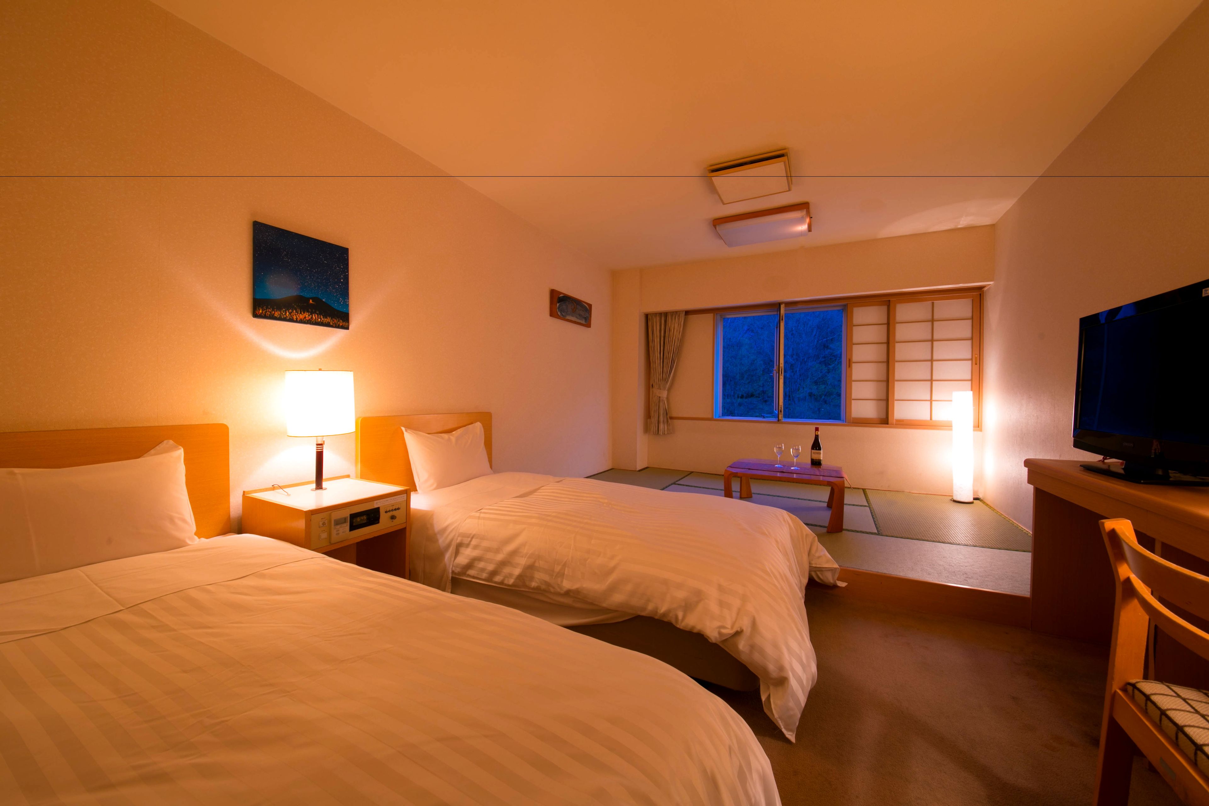 A hotel room with two beds, a nightstand with a lamp, a small table with a bottle, a television, and a window with curtains.
