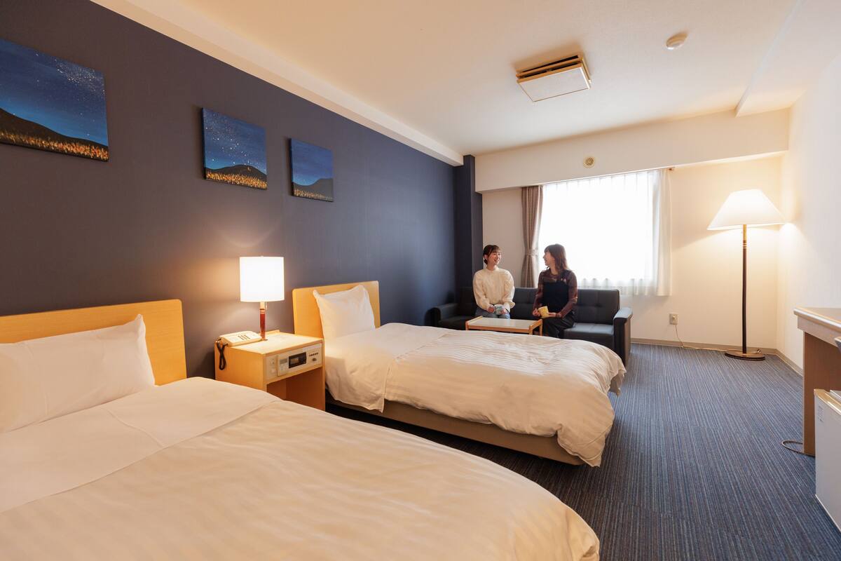 Une chambre d’hôtel avec deux lits, une table de chevet avec une lampe, un bureau et une chaise. Il y a deux tableaux accrochés au mur.