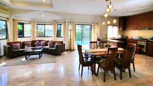 Amplia sala de estar con sofá, mesa de comedor y sillas, cocina con armarios de madera y ventanas grandes con vistas al exterior.