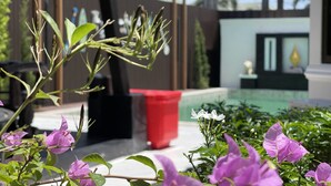 Zona de piscina con un bote de basura rojo, una silla blanca y flores púrpuras al frente.
