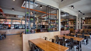 A modern restaurant interior with wooden tables and metal chairs, a counter with a menu, and shelves with plants and kitchen equipment. - MBI Desaku Homestay (Karangan)