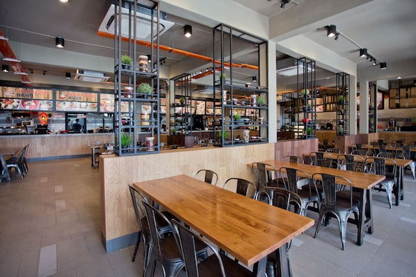 Ein modernes Restaurant-Interieur mit Holztischen und Metallstühlen, einer Theke mit Speisekarte sowie Regalen mit Pflanzen und Küchengeräten.