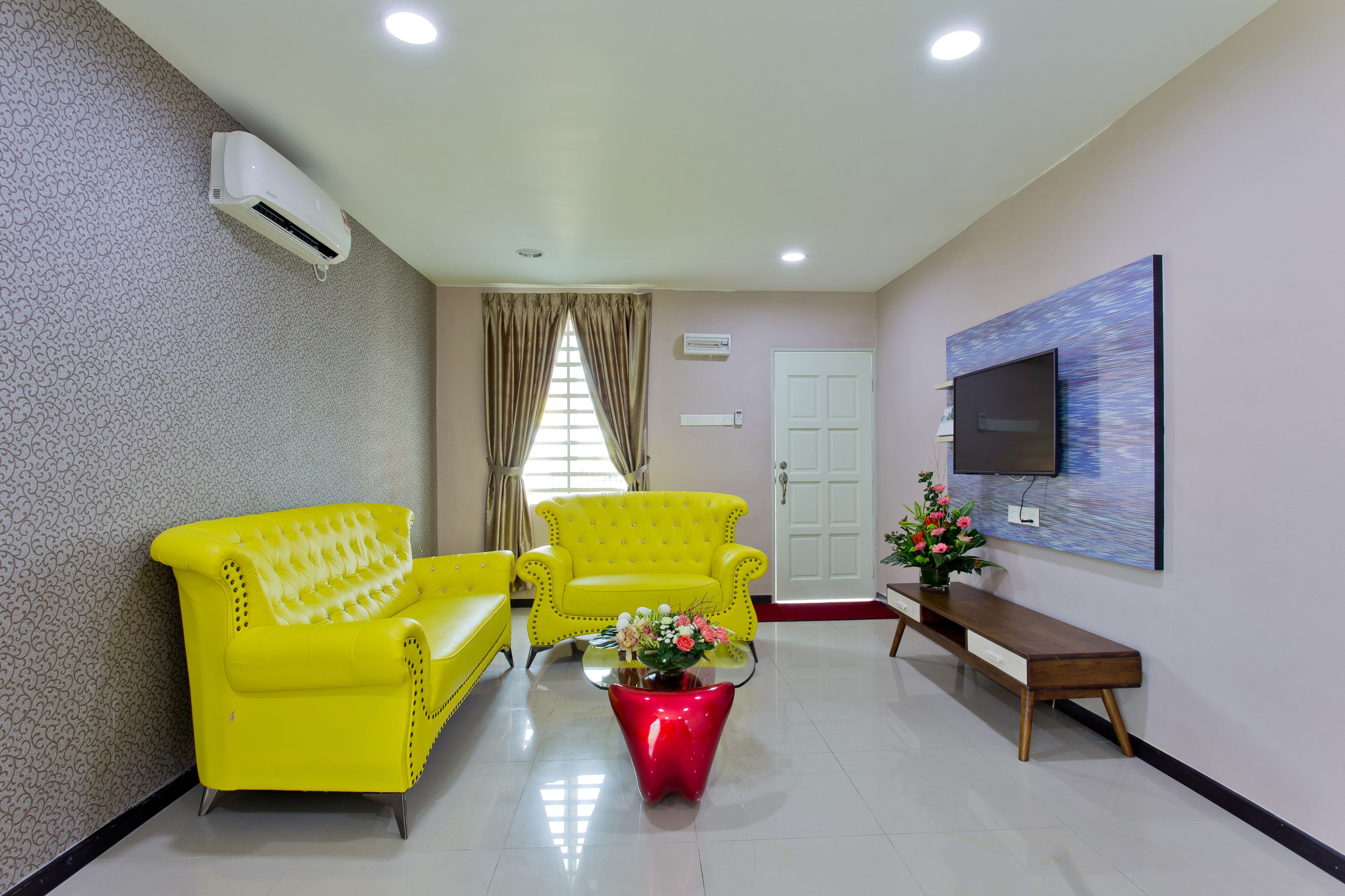 A room with two yellow armchairs, a red coffee table with flowers, a wooden bench, a TV on the wall, and a door.
