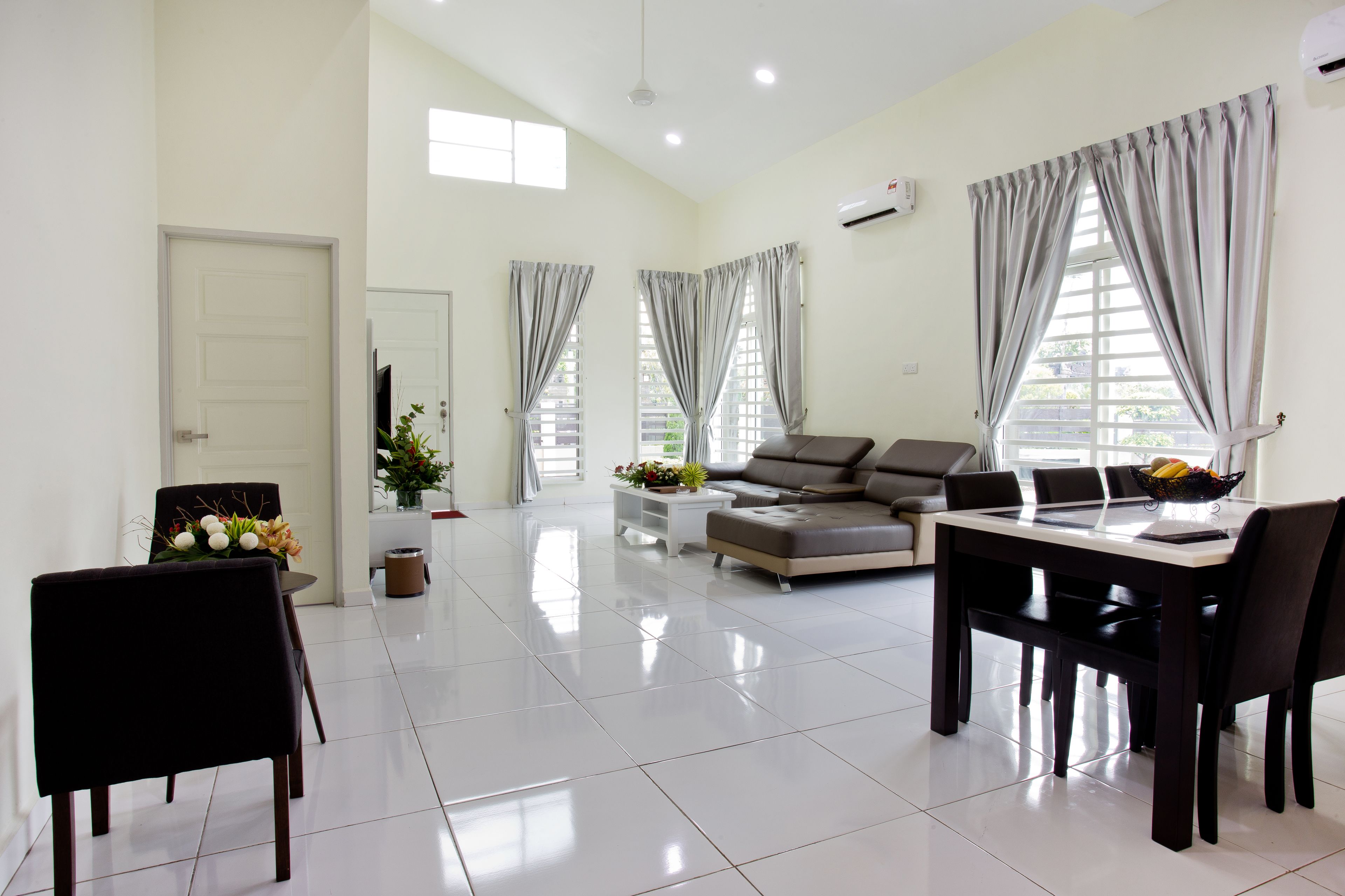 A spacious living room with a sofa, dining table, and chairs. There are large windows with curtains, a door, and a potted plant.
