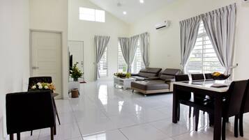 A spacious living room with a sofa, dining table, and chairs. There are large windows with curtains, a door, and a potted plant.