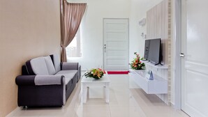 A modern living room with a sofa, a coffee table with a floral centerpiece, a flat-screen TV on a white console, and a door with a red threshold. - MBI Desaku Homestay (Karangan)