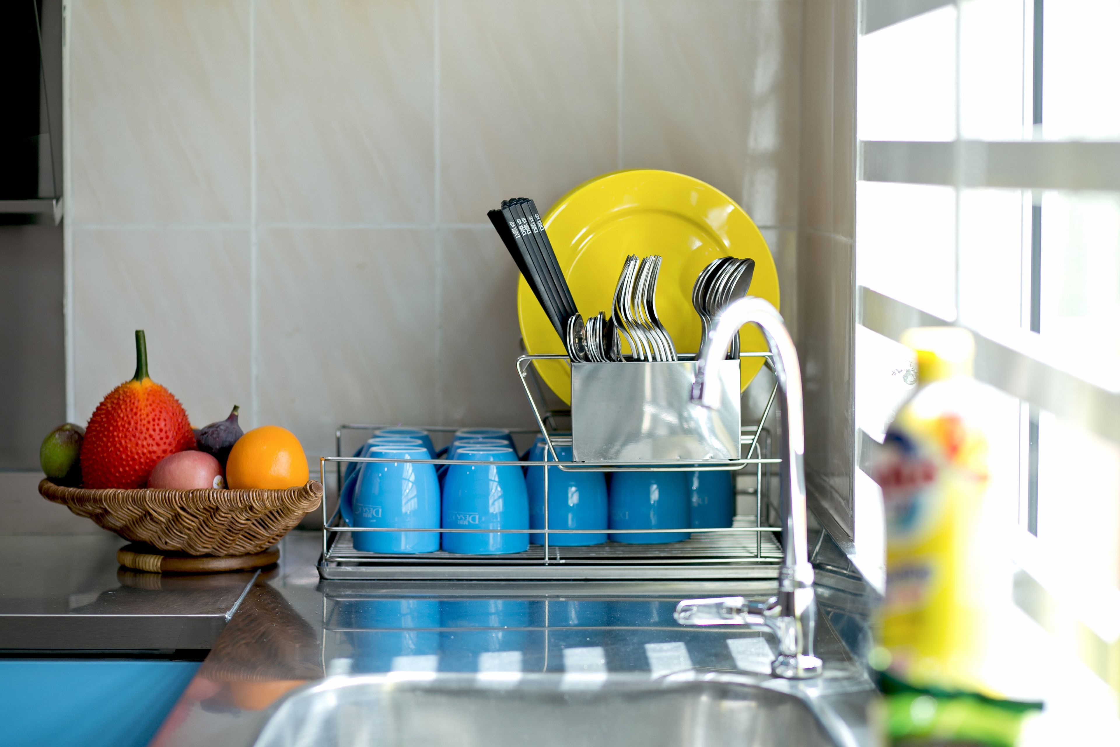 A kitchen with a sink, a dish rack with utensils, a basket of fruit, and a blue dish soap dispenser.